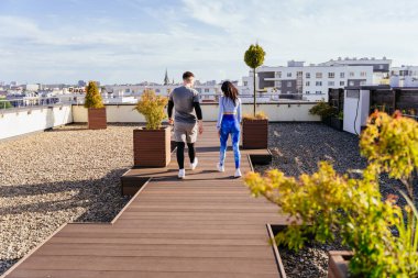 Arkadaşların yazın terasta buluşup şehre bakıyorlar. Erkek ve kadın çatıda egzersiz yapıyor ve dinleniyor. geniş açı arka görünümü.