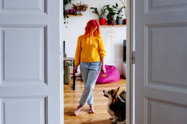 Young caucasian woman with vibrant orange hair in casual yellow sweater and jeans stands in cozy home interior. Corgi dog looks up at her with affection. Companionship, and a creative spring vibe. clipart