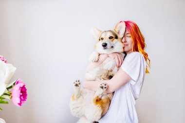 Renkli saçlı enerjik genç kadın beyaz arka planda sevimli Corgi köpeğini kucaklıyor. Çiçekler, sıcaklık ve mutlulukla çevrili neşeli, sevgi dolu bir an. İnsan ve hayvan, aşk, neşeli yaşam.