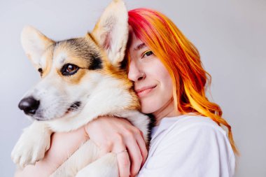 Canlı turuncu ve kızıl saçlı genç bir kadın beyaz arka planda Corgi köpeğini kucaklıyor. İnsan ve evcil hayvan arasındaki neşeli bağ, mutluluk hissi. Arkadaşlık ve renkli yaşam tarzı.