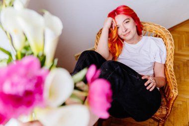 Canlı kızıl saçlı beyaz kadın hasır koltukta rahat bir şekilde oturuyor ve hafifçe gülümsüyor. Yumuşak çiçeklerle çevrili huzurlu bir atmosfer. İç gözlem ev sakinliği, günlük atmosfer.