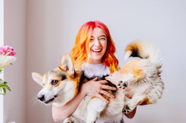 Turuncu ve pembe saçlı genç beyaz kadın tüylü bir Corgi köpeğini neşeyle içeride tutuyor. Arkadaşlık ve mutluluk. İç mekan, parlak doğal ışık, bir buket çiçek, hoş geldin havası..
