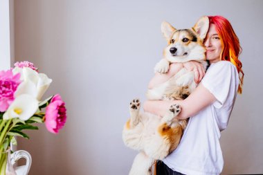 Renkli saçlı beyaz kadın tüylü Corgi köpeği tutuyor, canlı çiçeklerin yanında mutlu bir şekilde gülümsüyor. Sıcaklık, şefkat, arkadaşlık, neşeli atmosfer, insan ve evcil hayvan arasındaki bağ..