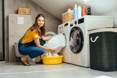 30 'lu yaşlarda bir kadın modern çamaşırhanenin içinde çamaşır makinesini gösteriyordu. Temiz çevre, günlük ev işleri, bakım ve organizasyon rutini.
