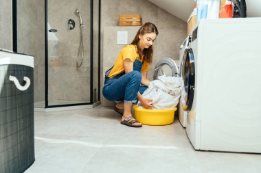 Çamaşırları yıkamadan önce düzenliyorum. Genç ve mutlu bir ev hanımı, modern, minimalist bir iç mekanda çamaşır makinesine çarşaf atar ya da çıkarır..