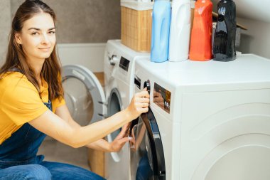 Güzel genç kadın ev işi yapıyor, çamaşır makinesini evde yıkıyor. Günlük ev hayatı, verimlilik ve rutin faaliyetleri şık ve işlevsel bir iç mekanda vurguluyor.