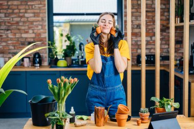 Profesyonel, sevimli kadın, evde bahçeyle uğraşmaktan zevk alıyor. Kaktüslü ayak parmakları, modern, rahat ev arka planında rüya gibi bir yüz ifadesi olan yanaklarının yanında küçük saksılar tutuyor..