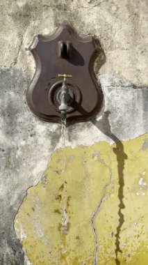 Bir dökme demir çeşmenin videosu çatlak sarı ve gri bir duvarın üzerine su dökerek çarpıcı bir görsel karşıtlık oluşturur.. 