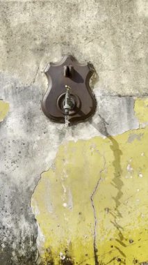 Bir dökme demir çeşmenin videosu çatlak sarı ve gri bir duvarın üzerine su dökerek çarpıcı bir görsel karşıtlık oluşturur.. 