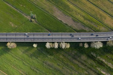 Yeşil kırsal kesimden geçen yoğun bir otoyolun hava manzarası, hareket halindeki arabalar ve yan taraflarında ağaçlarla. 