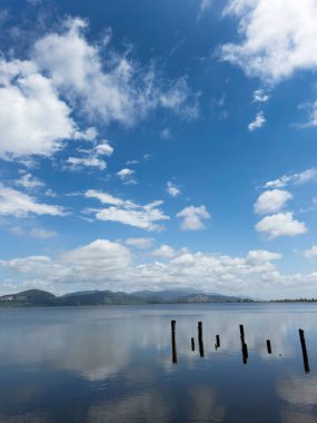 Göl yüzeyinin durgunluğunun fotografik belgeleri, beyaz bulutlarla benekli mavi bir gökyüzünü, arka planda dağları, Torre del Lago 'nun manzarasını yansıtır. 