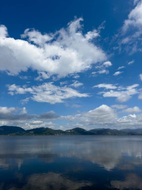 Göl yüzeyinin durgunluğunun fotografik belgeleri, beyaz bulutlarla benekli mavi bir gökyüzünü, arka planda dağları, Torre del Lago 'nun manzarasını yansıtır. 
