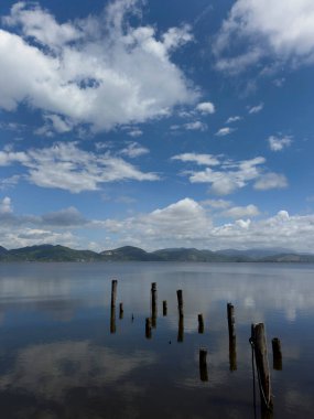 Göl yüzeyinin durgunluğunun fotografik belgeleri, beyaz bulutlarla benekli mavi bir gökyüzünü, arka planda dağları, Torre del Lago 'nun manzarasını yansıtır. 