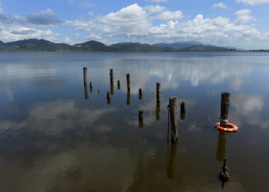 Göl yüzeyinin durgunluğunun fotografik belgeleri, beyaz bulutlarla benekli mavi bir gökyüzünü, arka planda dağları, Torre del Lago 'nun manzarasını yansıtır. 