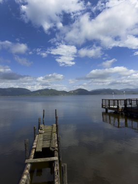 Göl yüzeyinin durgunluğunun fotografik belgeleri, beyaz bulutlarla benekli mavi bir gökyüzünü, arka planda dağları, Torre del Lago 'nun manzarasını yansıtır. 