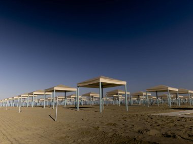 Güneşli bir günde kumlu ve tırmıklı bir plajda dizilmiş beyaz ve mavi şemsiyelerin görüntüsü.