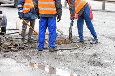 Yol işçilerinden oluşan bir ekip eski asfaltı kanalizasyon boşluğunun etrafından bir kaya matkabı ve kürekle temizliyor. Boşluğu kopyala.