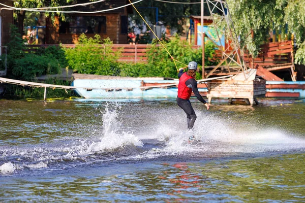 An athlete wakeboarder cuts the water surface at high speed against a blurred background of the river bank creating a cloud of splashes around him.