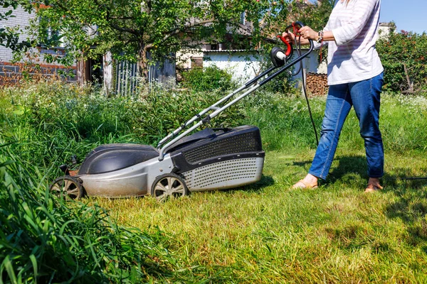 女性の庭師は庭の背の高い緑の草を切ります 庭師は背の高い草を裏庭に電動芝刈り機で刈る — ストック写真