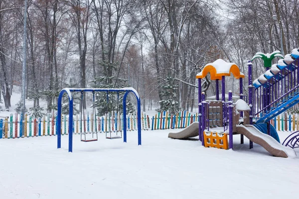 Winter landscape of a snow-covered playground in a city park after a snowfall. Multi-colored slides and metal swings are sprinkled with fluffy snow. Copy space.