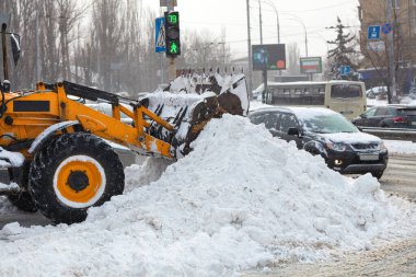 The blade of a communal tractor clears piles of snow from a city street among the carriageway on a winter day during a snowfall.