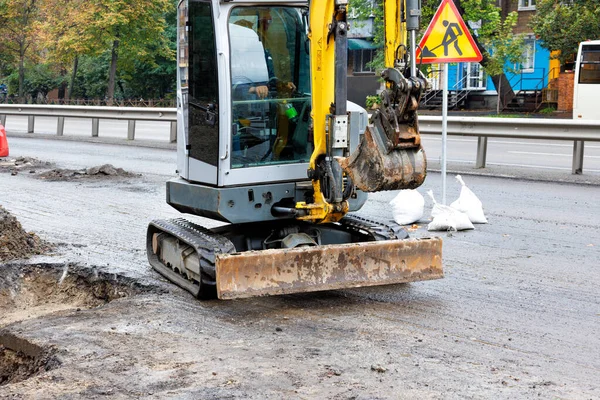 stock image A road mobile excavator digs a roadbed for the repair of sewer infrastructure on the carriageway of a city street. Copy space.