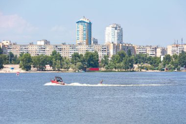 Summer river landscape with a motor boat and water skis on the river against the backdrop of the urban landscape and residential buildings.