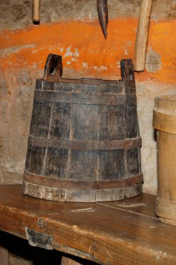 An old wooden bucket, upholstered around the circumference with forged metal hoops, stands on a wooden bench in the corner of a village hut. Vertical image.