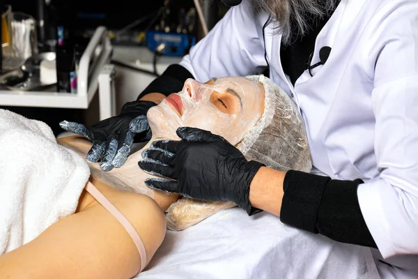 The hands of a beautician apply a cleansing cream on the face of a young woman in the process of a facial cleansing procedure.