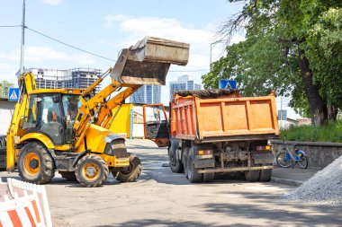 Güneşli bir günde eski bir kamyonun arkasına çakıl yüklemeden önce yol üzerinde büyük bir hidrolik çatallı kova taşıyan bir traktör. Boşluğu kopyala.