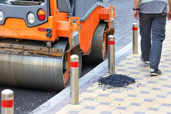 The road vibratory roller with its metal wheels compacts fresh asphalt with high precision along the edge of the road along the paved sidewalk.