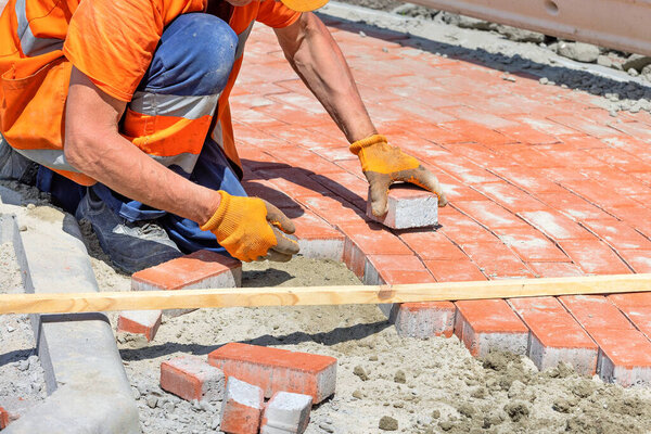 A bricklayer in work gloves and overalls lays red paving slabs on a sunny day. 05.22.2023. Kyiv.Ukraine.