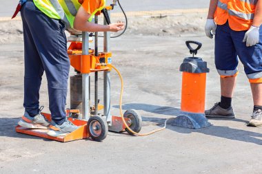 Yansıtıcı pelerinli yol işçileri benzin sondaj makinelerini güneşli bir günde iş yerlerindeki kalınlığını ölçmek için asfaltı kesecek şekilde ayarlıyorlar. Boşluğu kopyala.