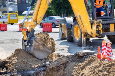 Bir inşaat kazıcı operatörü, güneşli bir yaz gününde bir şehir caddesinin zeminine hendek kazmak ve kum boşaltmak için ustalıkla bir bumbar ve kova kullanıyor. Boşluğu kopyala.