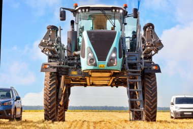 A high wheelbase industrial sprayer for the agricultural sector stands in comparison with passenger cars in a harvested field against a blue cloudy sky. clipart