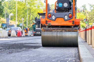 Titreşen bir patenin metal silindiri güneşli bir günde kaldırım kenarındaki yolda şehir sokağının arka planına karşı taze asfaltı sıkıştırır. Yakın plan. Boşluğu kopyala.
