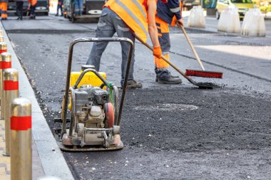 Yaya kaldırımının kenarındaki taze asfalt üzerinde yıpranmış bir levha bulanık bir şekilde çalışan bir yol işçisinin arka planına karşı duruyor..