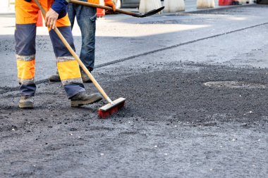 Yol işçileri kanalizasyon kanallarının etrafındaki yeni asfalt yüzeyinde bir fırça ve kürekle fazla moloz kaldırıyorlar. Boşluğu kopyala.