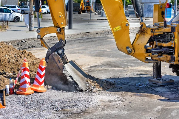 stock image The bucket of a large construction excavator skillfully lifts a layer of old asphalt to dig a trench on a job site against the backdrop of a city street on a sunny summer day. Copy space.