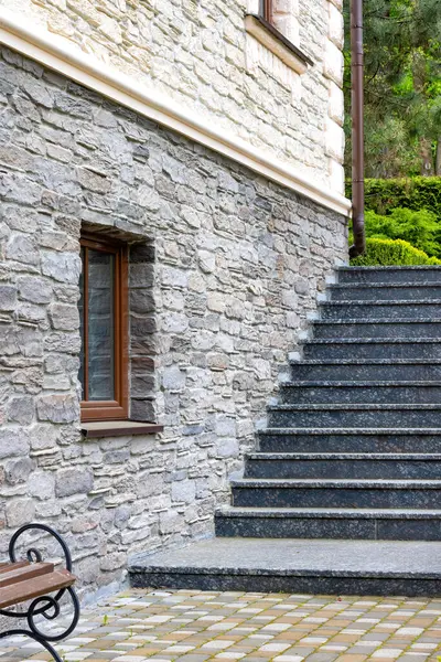 Granite steps along the stone wall of the house with a window lead to the green garden in the background. Vertical image.