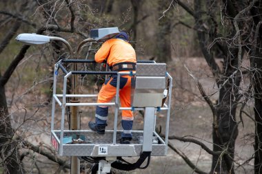 Parlak turuncu bir güvenlik elbisesi içinde, yüksek bir platformda çalışan işçiler sokak lambasını tamir ediyorlar. Etrafı çıplak ağaçlarla çevrili, bakım işi, dış mekan konsepti.