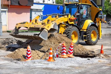 İnşaat konileri ve molozlarla çevrili büyük sarı bir kepçe kazıcısı toprağı kazarken, işçiler kalabalık bir kentsel alandaki sokakları onarmaya odaklanıyor..