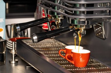 Pürüzsüz bir espresso makinesi kullanarak canlı turuncu bardaklarda iki tane espresso hazırlıyor. Kahve kokusu havayı doldururken kafe enerji ile uğulduyor..