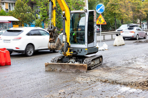Компактный экскаватор старательно работает, чтобы сгладить мокрую дорогу среди городского движения, с красочными автомобилями, проходящими мимо и знаками, направляющими водителей, все под облачным небом.