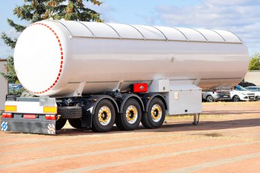 A large white fuel tanker stands on a brick paved area, showcasing its shiny surface under the sunlight. The cargo vehicle is surrounded by an industrial backdrop and a few cars nearby. clipart