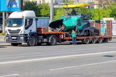 Büyük bir kamyon düz bir karavanda yeşil bir yol yapım makinesi taşıyor. Bir işçi, açık mavi gökyüzünün altındaki işlek bir şehir sokağındaki yükleme işlemini izliyor ve gün ortası etkinliğini gösteriyor.