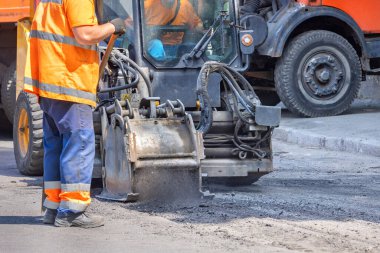 Turuncu güvenlikli yelek giyen bir inşaat işçisi özenle kompakt bir yol tamir makinesini çalıştırıyor. İşçi yolda asfaltı düzleştirirken gün ışığı sahneyi aydınlatıyor..