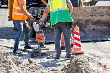 Güvenlik yelekli iki inşaat işçisi bir yol onarım alanında toprağı sıkıştırmak için ağır makineler kullanıyor. Parlak turuncu koniler açık mavi gökyüzünün altındaki çalışma alanının aktif bir atmosfer yarattığını gösteriyor..