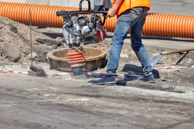 Turuncu yelekli bir inşaat işçisi yoğun bir caddede zemini sıkıştırmak için özel ekipman kullanır. Etrafındaki enkaz ve turuncu borular devam eden altyapı çalışmalarını gösteriyor. Boşluğu kopyala.