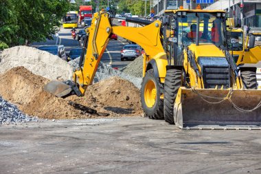 Toprak taşıyıcılar ve yükleyiciler özenle çalışarak şehri yeniden şekillendiriyorlar. İşçiler yoğun trafiğin ortasında, öğlen güneşinin altında kum ve çakıl yığınlarını yönetirken havada toz dansları yapılıyor. Boşluğu kopyala.